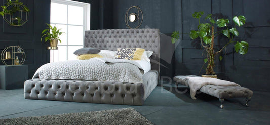 Gray tufted bed in a modern bedroom with plants and a rug.